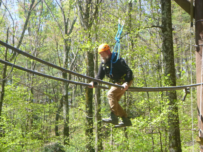 A man wearing a helmet and safety harness is carefully walking across a rope bridge high up in the trees. He is holding onto the ropes for balance as he navigates the challenging obstacle. The surrounding forest is lush and green, with sunlight filtering through the leaves. The scene captures a moment of adventure and focus as the man tackles the ropes course.